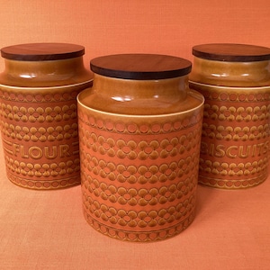 Op de afbeelding: Drie oranje keramische keukencanners met houten deksels. De potten hebben een herhalend bloemmotief en de woorden "FLOUR" en "BISCUITS". De deksels zijn donkerbruin.