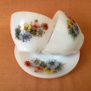 May include: Two white ceramic teacups with floral designs in shades of blue, yellow, and red. The cups are stacked on top of each other and are sitting on a white saucer with a matching floral design.