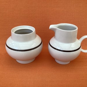 May include: A white ceramic sugar bowl and creamer set with a black stripe around the middle. The set is on a textured orange surface.