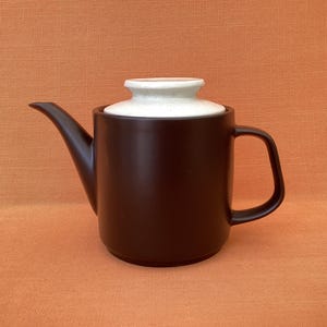 Könnte beinhalten: Dunkelbraune Keramik-Teekanne mit passendem Griff und Ausguss. Der Deckel ist hellbeige. Die Teekanne steht auf einer orangefarbenen Oberfläche. Perfekt zum Aufbrühen von Tee.