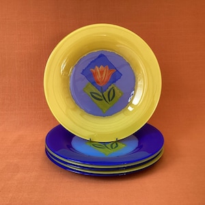 May include: A set of decorative plates featuring a floral design. The plates have a yellow rim and a blue centre with a red and orange tulip design. The plates are stacked on an orange surface.