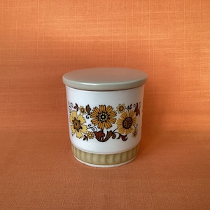 Könnte beinhalten: Eine weiße Keramikdose mit einem Blumendesign in Braun-, Gelb- und Orangetönen. Der Deckel ist hellgrau.