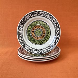 May include: A stack of three white ceramic plates featuring a decorative pattern. The rim has a black swirling scroll design. The centre displays a green circular band surrounding a brown and green floral motif with additional swirling details.