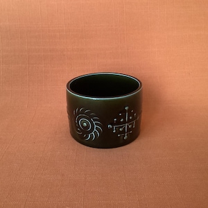 Può includere: Una tazza in ceramica verde scuro con disegni incisi bianchi. La tazza presenta una spirale a forma di sole e un motivo a forma di croce. La tazza è cilindrica e poggia su una superficie arancione-marrone.