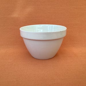 May include: A solid white ceramic mixing bowl with a smooth, matte finish and a wide, circular rim. The bowl features a deep, rounded basin that tapers toward a smaller, flat base, providing a stable structure for various kitchen tasks.