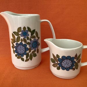May include: Two white ceramic pitchers with floral designs. The larger pitcher has a tall, curved handle and a spout. The smaller pitcher has a shorter handle. Both have blue and green floral patterns.