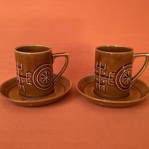 May include: Two brown ceramic coffee cups with matching saucers. Each cup has a handle and is decorated with a white geometric design and a starburst. The cups and saucers are on an orange background.