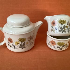 Könnte beinhalten: Ein weißes Keramik-Teeservice mit einer Teekanne mit Deckel, einem Sahnekännchen und einer Zuckerdose. Das Set ist mit einem floralen Muster in Orange-, Gelb- und Grüntönen verziert. Die Teekanne hat einen gebogenen Griff und Ausguss.