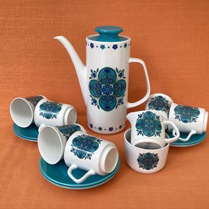 May include: A vintage white ceramic coffee set with a teal floral design. The set includes a tall coffee pot, six cups with saucers, and a creamer. The coffee pot has a teal lid and handle. The cups and creamer have a matching teal floral pattern.