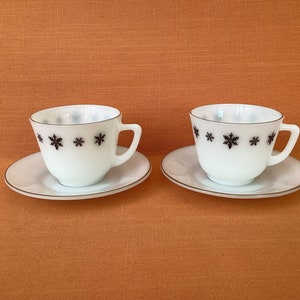 May include: Two white teacups with black snowflake designs and matching saucers. The cups and saucers have a gold rim.