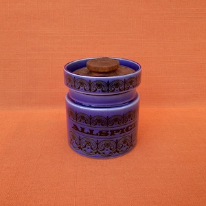 Può includere: Un barattolo di spezie in ceramica blu con coperchio in legno, etichettato "ALLSPICE" in nero. Il barattolo presenta decorazioni a volute nere sulla parte superiore e inferiore. Lo sfondo è arancione.