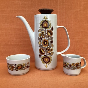 May include: A white ceramic coffee pot with a brown lid and a floral pattern in shades of brown, orange, and yellow. The pot is accompanied by two matching sugar and creamer cups.