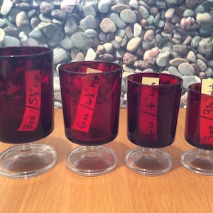 Peut inclure: Quatre bougeoirs en verre rouge avec des bases en verre transparent. Les bougeoirs sont étiquetés avec leur taille et leur hauteur : 8 oz / 5 1/2" de haut, 6 oz / 4 1/2" de haut, 4 oz / 4" de haut et 3 oz / 3 1/2" de haut.