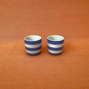May include: Two small, cylindrical ceramic cups featuring a repeating horizontal striped pattern in alternating bands of deep blue and white. Each cup has a smooth, rounded rim and a white interior, standing side by side as a matching pair.
