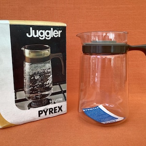 May include: A clear glass Pyrex Juggler jug with a black handle and lid, next to its original box. The box features the product name and an image of the jug. The jug is designed to hold 1.9 litres.