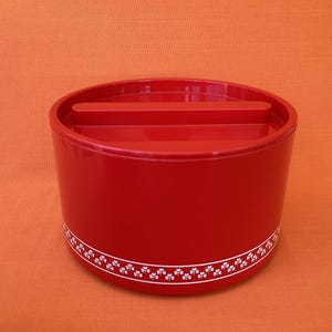 May include: A red, cylindrical container with a matching lid. The container has a white floral pattern around the base. The background is a solid orange colour. The container is likely for kitchen or home storage.