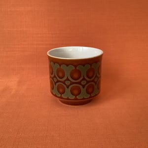 May include: A ceramic mug with a brown exterior and a white interior. The mug features a repeating pattern of brown and green circles. The mug is set against an orange background.