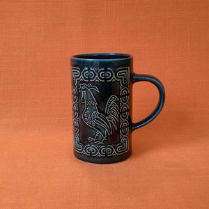 May include: A dark blue ceramic mug with a white rooster design. The rooster is surrounded by a geometric pattern.
