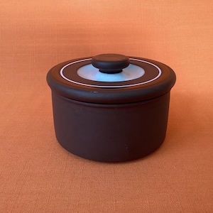 May include: A small, round, dark brown ceramic container with a matching lid. The lid has a light blue centre with a white ring and a small, rounded knob. The container sits on an orange surface.