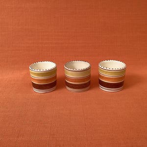 May include: Three small ceramic cups featuring horizontal stripes in shades of yellow, orange, and dark brown. Each cup has a white interior and a rim decorated with a series of small, evenly spaced dark brown dots around the top edge.