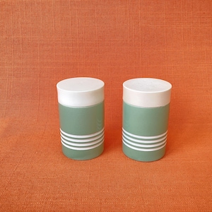 May include: Two matching salt and pepper shakers. Each shaker has a white lid and a light green body with three white horizontal stripes. The shakers are cylindrical and sit on an orange surface.