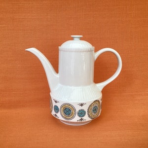 Könnte beinhalten: Weiße Kaffeekanne aus Keramik mit einem geometrischen Muster in Blau und Gold. Die Kanne hat einen runden Korpus und einen Ausguss auf einer Seite.