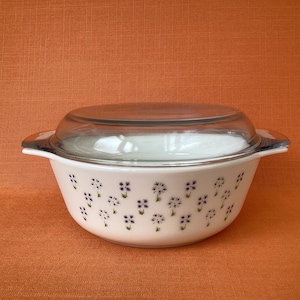 Könnte beinhalten: Weiße Auflaufform mit Glasdeckel. Die Form hat ein Blumenmuster mit violetten Blüten und grünen Stielen.