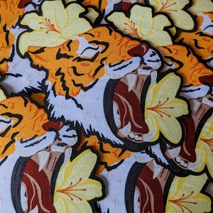 May include: Close-up of several embroidered patches featuring a roaring tiger with a yellow flower behind it. The tiger is orange and black with a white background. The flower is yellow with a white center.
