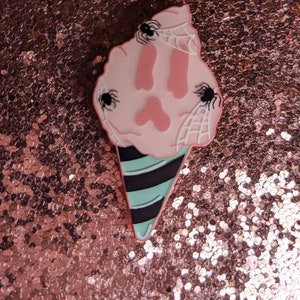 May include: A pink and white ice cream cone with black spiders and cobwebs. The cone is striped with black and green.