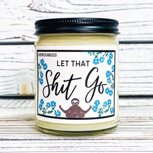 May include: A white candle jar with a black lid and a label that says "Let That Shit Go" with a cartoon sloth meditating in the center. The label has a blue floral pattern.