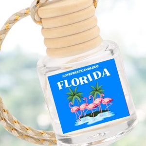 May include: A clear glass car air freshener with a wooden cap and a braided rope. The label features the word "FLORIDA" in white letters on a blue background, along with palm trees and pink flamingos. The text "LOVEFIRSTCANDLECO" is above the word "FLORIDA".
