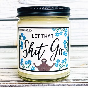 May include: A white candle in a glass jar with a black lid. The label on the jar has a blue floral design and says "Let That Shit Go" with a cartoon sloth in a yoga pose.