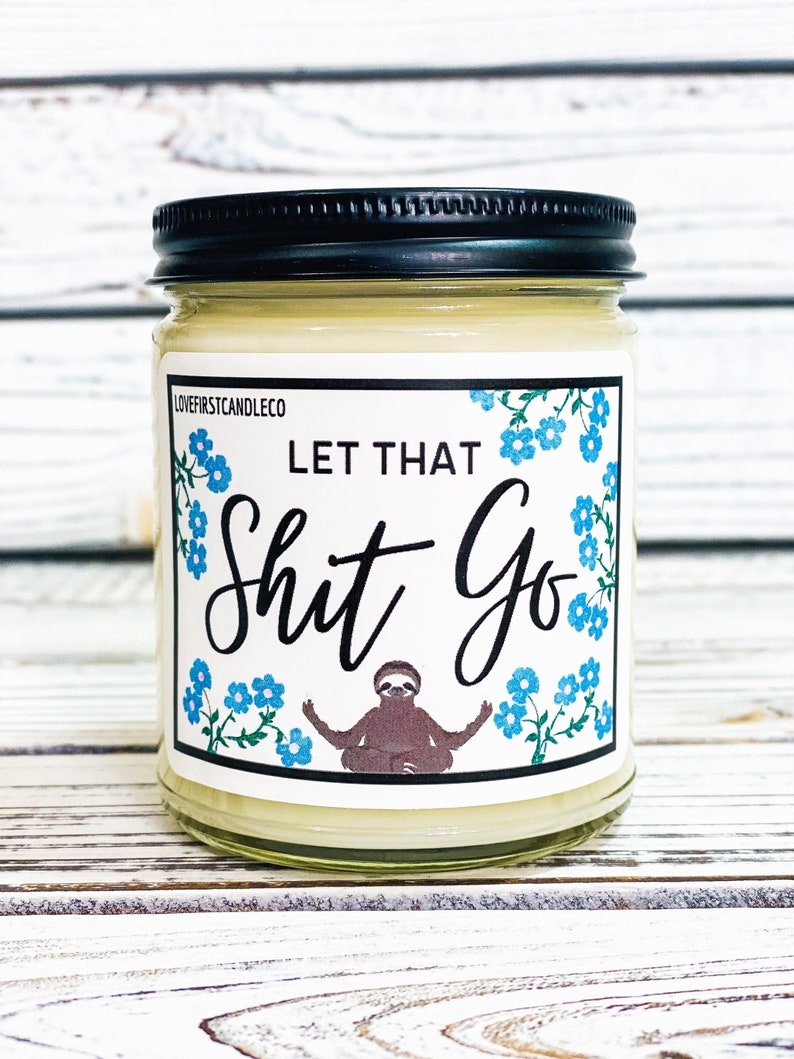 May include: A white candle in a glass jar with a black lid. The label on the jar has a blue floral design and says "Let That Shit Go" with a cartoon sloth in a yoga pose.