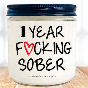 May include: A white candle jar with a black lid. The jar has a red heart and black text that reads "1 YEAR F*CKING SOBER" and "LOVEFIRSTCANDLECO".