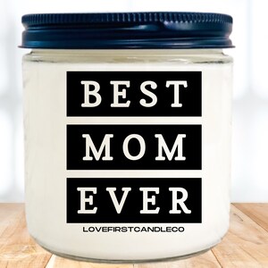 May include: A white candle jar with a black lid. The jar has black text that reads "BEST MOM EVER" and the text "LOVEFIRSTCANDLECO" at the bottom.