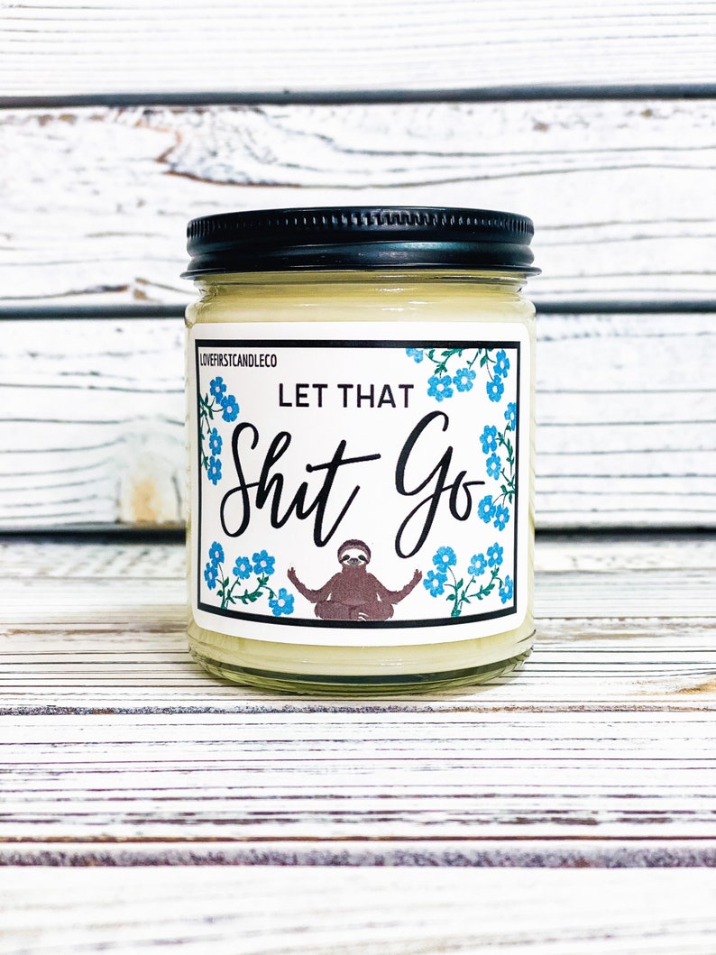 May include: A white candle jar with a black lid and a label that says "Let That Shit Go" with a cartoon sloth meditating in the center. The label has a blue floral pattern.