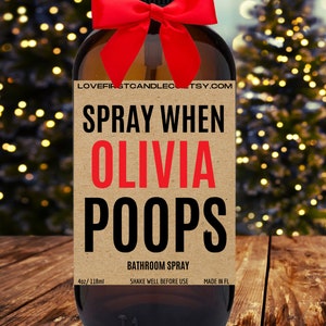 May include: A brown glass bottle with a red bow and a label that says "SPRAY WHEN OLIVIA POOPS" in black and red text. The label also says "BATHROOM SPRAY", "4oz/118ml", "SHAKE WELL BEFORE USE", and "MADE IN FL".