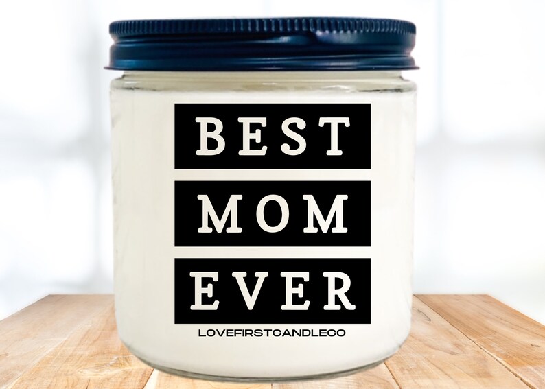 May include: A white candle jar with a black lid. The jar has black text that reads "BEST MOM EVER" and the text "LOVEFIRSTCANDLECO" at the bottom.