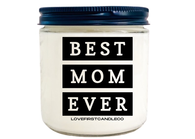 May include: A white candle jar with a black lid. The jar has a black label with the text "BEST MOM EVER" in white letters. The label also includes the text "LOVEFIRSTCANDLECO" in smaller white letters.