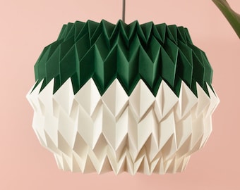 Könnte beinhalten: Eine weiße und grüne Pendelleuchte im Origami-Stil hängt an der Decke vor einer rosa Wand. Die Leuchte ist aus Papier gefertigt und hat ein geometrisches Design. Eine klare Glasvase mit strukturierter Oberfläche steht auf einem weißen Sockel. Die Vase enthält eine grüne, belaubte Pflanze.