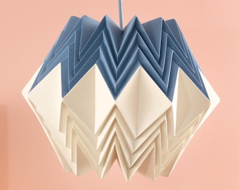 Könnte beinhalten: Ein weißer und blauer Origami-Papierlampenschirm hängt an einem Kabel vor einem rosa Hintergrund. Der Lampenschirm besteht aus gefaltetem Papier und hat ein geometrisches Design. Im Vordergrund befinden sich Holzblöcke auf einem rosa Sockel.