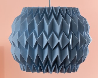 Könnte beinhalten: Eine blaue Hängelampe im Origami-Stil hängt von der Decke über einer rosa Oberfläche. Holzblöcke in verschiedenen Größen sind auf einem rosa Sockel angeordnet.
