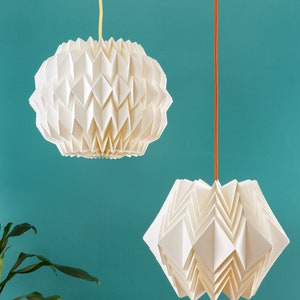 Puede incluir: Dos l&aacute;mparas colgantes de papel origami blanco que cuelgan del techo contra una pared azul turquesa. La luz de la izquierda es redonda y la luz de la derecha tiene forma de estrella.