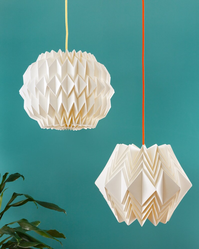 Puede incluir: Dos l&aacute;mparas colgantes de papel origami blanco que cuelgan del techo contra una pared azul turquesa. La luz de la izquierda es redonda y la luz de la derecha tiene forma de estrella.