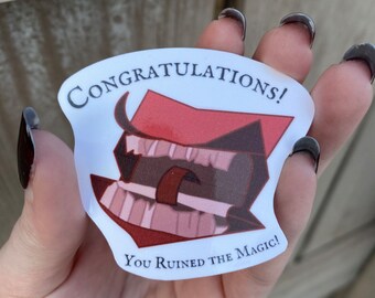 May include: A sticker with the text "Congratulations! You Ruined the Magic!" The sticker features a cartoon image of a red hat and a mouth with teeth. The sticker is held in a hand.