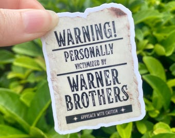 May include: A rectangular sticker with a distressed paper effect. The sticker features the text "WARNING! PERSONALLY VICTIMIZED BY WARNER BROTHERS" in bold, black font. The bottom of the sticker reads "APPROACH WITH CAUTION" with a star symbol.