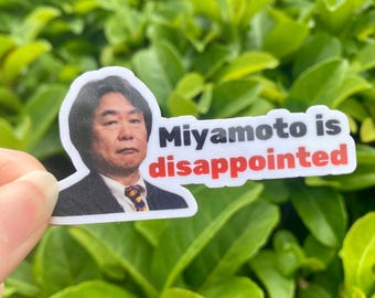 May include: A white sticker featuring a portrait of a man with the text "Miyamoto is disappointed" in black and red. The sticker is held against a backdrop of green foliage.