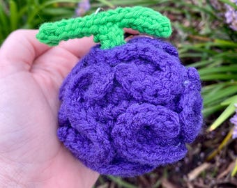 May include: A round, purple crocheted object with a textured, swirling surface pattern and a bright green crocheted stem attached to the top. The item is crafted from yarn and features a dense, knotted construction throughout its spherical body.