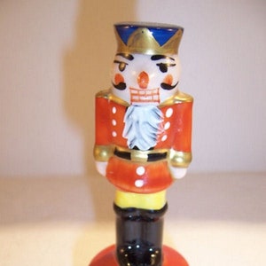 May include: A decorative ceramic nutcracker figurine. The figurine is painted with a red jacket, black boots, and a white face with a mustache. The nutcracker is standing on a red, round base with a hinged lid. The hat is blue and gold.