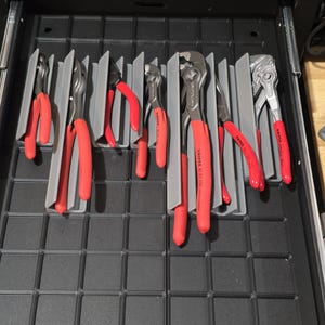 May include: A drawer organizer with various red-handled pliers and wrenches. The tools are neatly arranged in gray dividers within a black drawer. The tools are of different sizes and shapes, suggesting a set for various tasks.